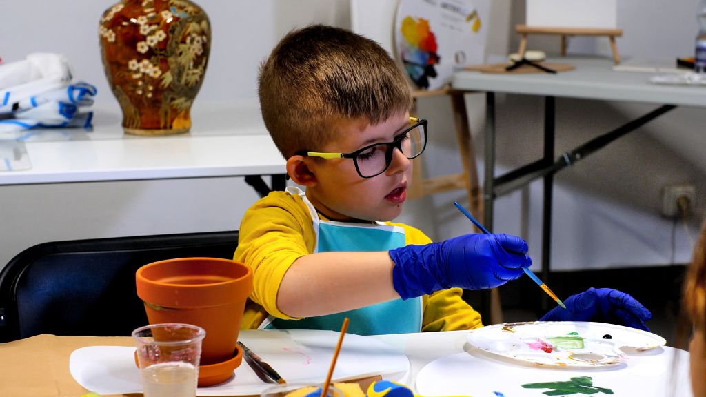 Kilkuletni chłopiec maluje farbami, ozdabia naczynie gliniane. Na stole przybory do malowania.