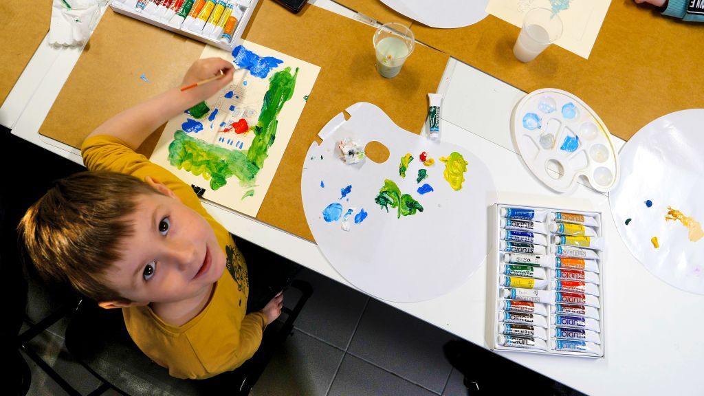 Kilkuletni chłopiec maluje obrazek. Na stole przybory do malowania (farby, pędzel, woda w kubeczkach, paleta do mieszania farb, kartony.
